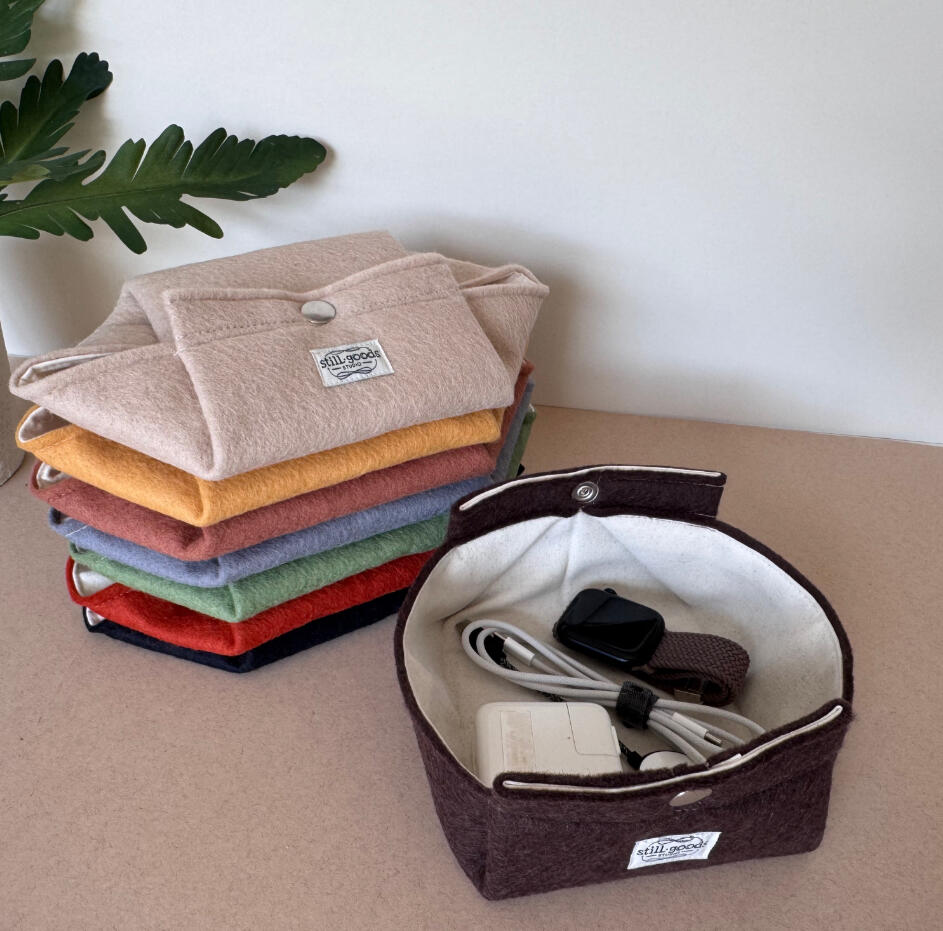 Foldable Travel Tray Stack of folded wool felt pouches in an array of colors, beside an open round felt caddy holding a charging cable, styled with a green plant leaf.