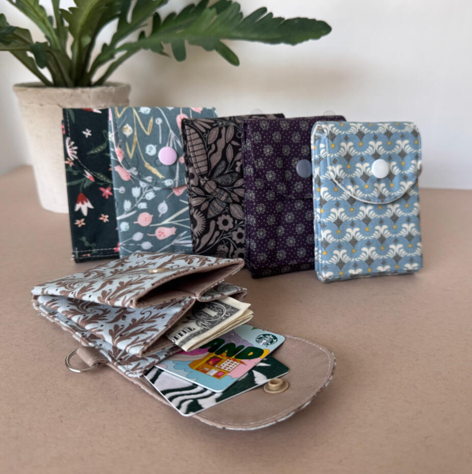 Accordion Wallet Snap-close fabric wallets in floral and botanical prints, opened to show cards tucked inside, styled with a small potted plant.